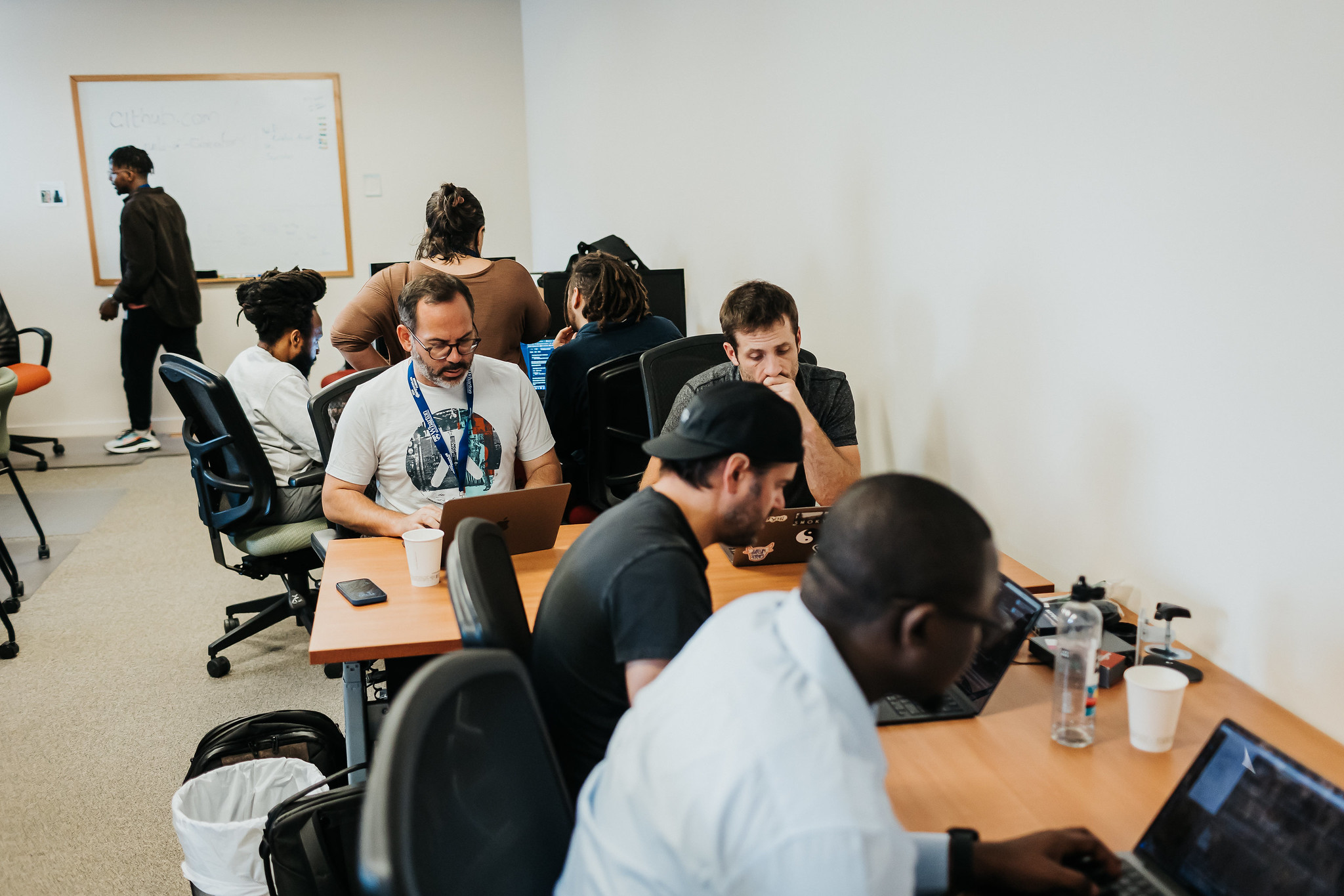 A photo of people at the sprints. Several people are at desks, looking intently at laptops. There is a group of three people discussing something in the background.