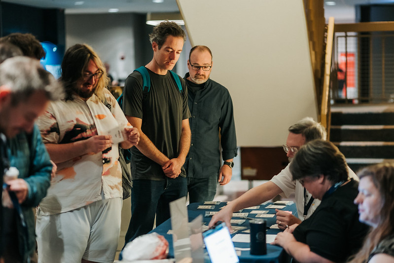 People gathered at an event, with attendees standing and talking while others sit at a table with name badges and event materials.